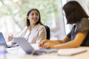 A woman sitting at a desk in a bright office space smiles while talking with a colleague. Both have laptops open in front of them, and large windows with blurred greenery outside are in the background.